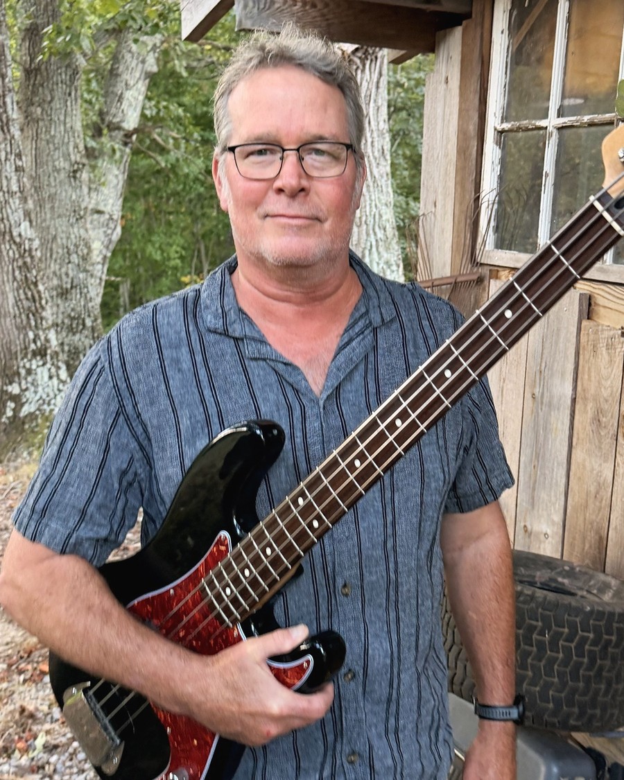 Brent standing by a weathered shed holding a black Fender Precision bass.