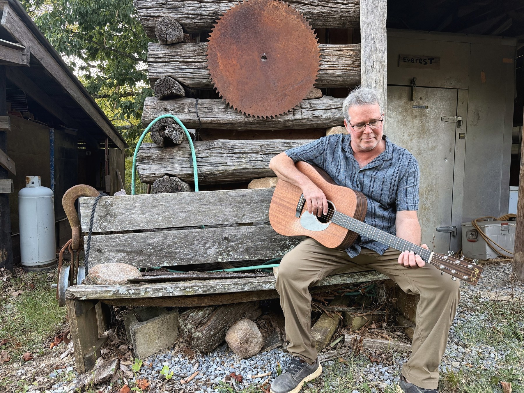 Brent sitting alone on the weathered bench outside the log outbuilding with the Martin across his knee, looking down at the strings, a rusted sawmill blade hung on the logs behind him.