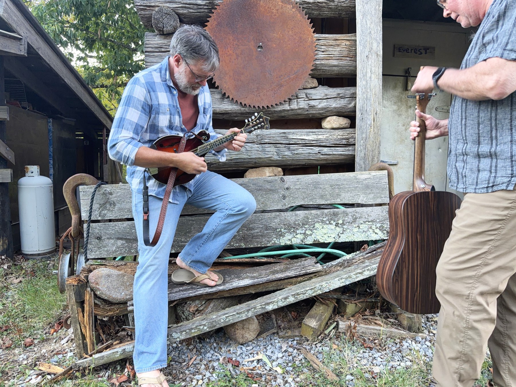 William playing the mandolin on a bench outside the log outbuilding while Brent stands beside him lifting a dreadnought out of its case.