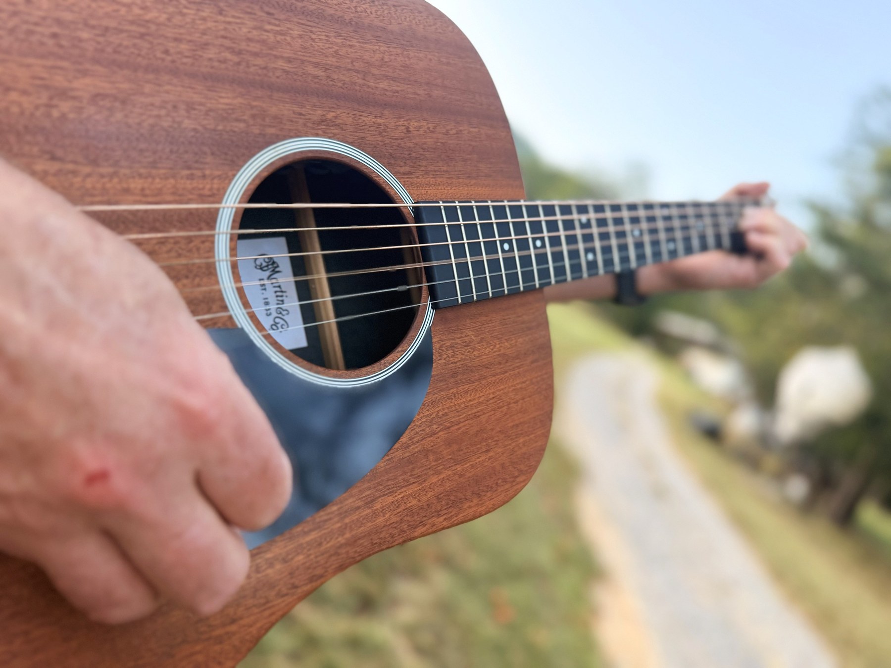 Close view across the soundhole of the mahogany Martin, strings catching the last of the light, a gravel farm track soft behind it.