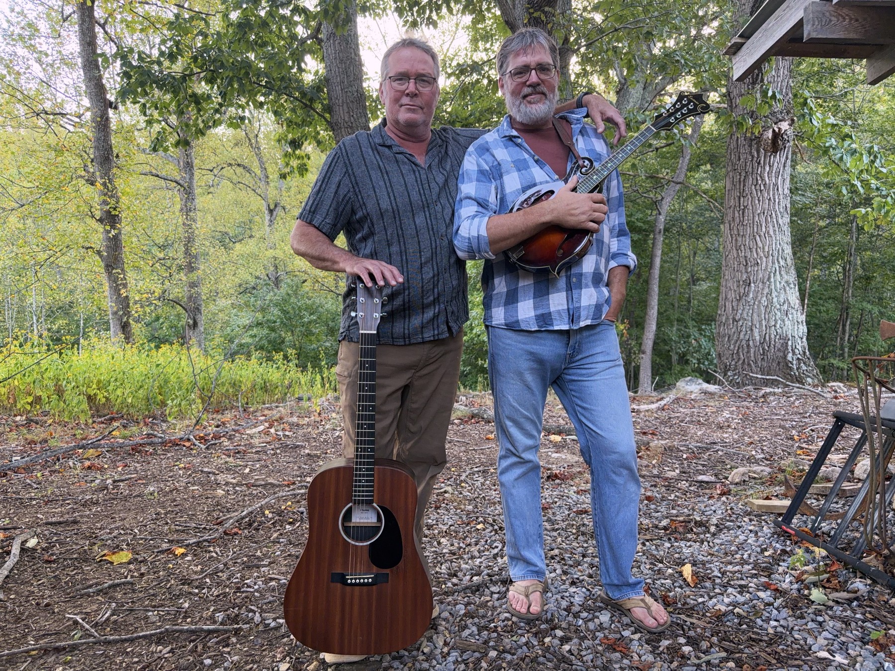 Brent and William standing side by side outside a log outbuilding at dusk, Brent's arm around William's shoulder, a Martin dreadnought stood on the ground and an Eastman mandolin held across the chest.