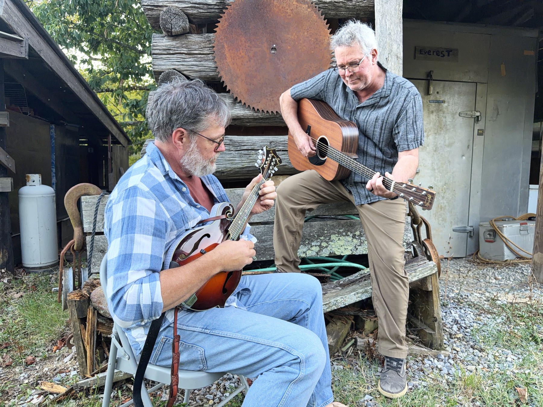 Brent and William playing outside the same outbuilding, William seated on a folding chair with the mandolin and Brent perched on a weathered bench with the guitar, a rusted sawmill blade hung on the logs behind them.